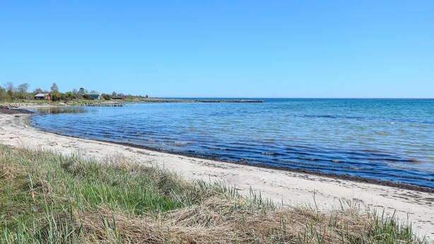 A beach with clear blue water, a sandy shore, and a few buildings in the background. - 3 bedroom gorgeous home in Löttorp (Löttorp)