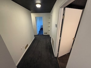Un couloir étroit, dont le sol est recouvert d’une moquette, avec une porte sur la droite et un luminaire au plafond.