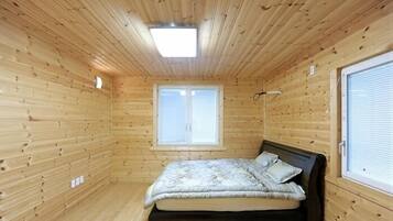 A small, wooden-paneled room with a bed, a window, and a ceiling light.