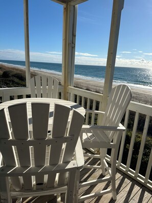 En täckt veranda med vita Adirondack-stolar med utsikt över en strand.