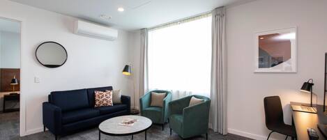 Una habitación de hotel moderna con un sofá azul oscuro, dos sillones verdes, una mesa de centro redonda y un escritorio con una computadora portátil.