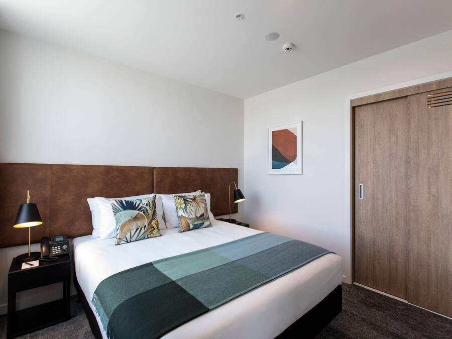 Habitación de hotel con una cama grande, cabecera de madera, mesita de noche con lámpara y un armario de madera.