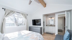 A bedroom with a bed, a grey sofa, a TV, and a kitchen area visible through an open door.