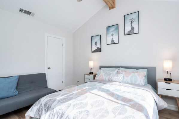 Quarto com cama, sofá cinza e três quadros na parede.