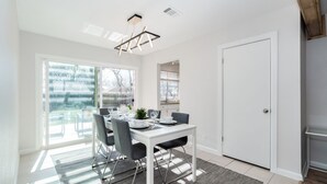 A modern dining area with a white table and chairs, a hanging light fixture, and a view of the outdoors through large windows.