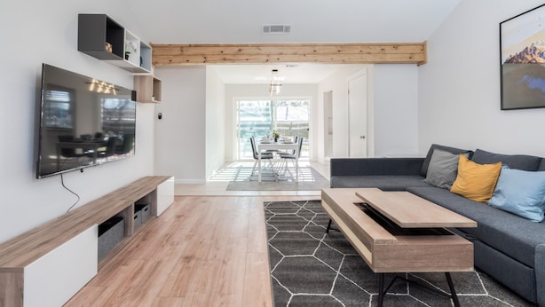 A modern living room with a flat-screen TV, wooden flooring, a gray sofa with cushions, and a dining area in the background.