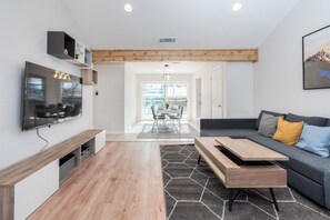 Uma sala de estar moderna com televisão de tela plana, piso de madeira, sofá cinza com almofadas e uma área de jantar ao fundo.