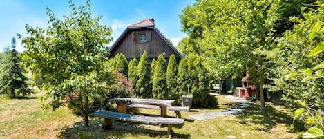 A rustic wooden building with a sloped roof, surrounded by lush greenery and a picnic table in the foreground.