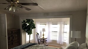 Ein Schlafzimmer mit Bett, Deckenventilator, Kommode, Stuhl, Tisch, Lampe und einem großen Fenster mit Vorhängen.