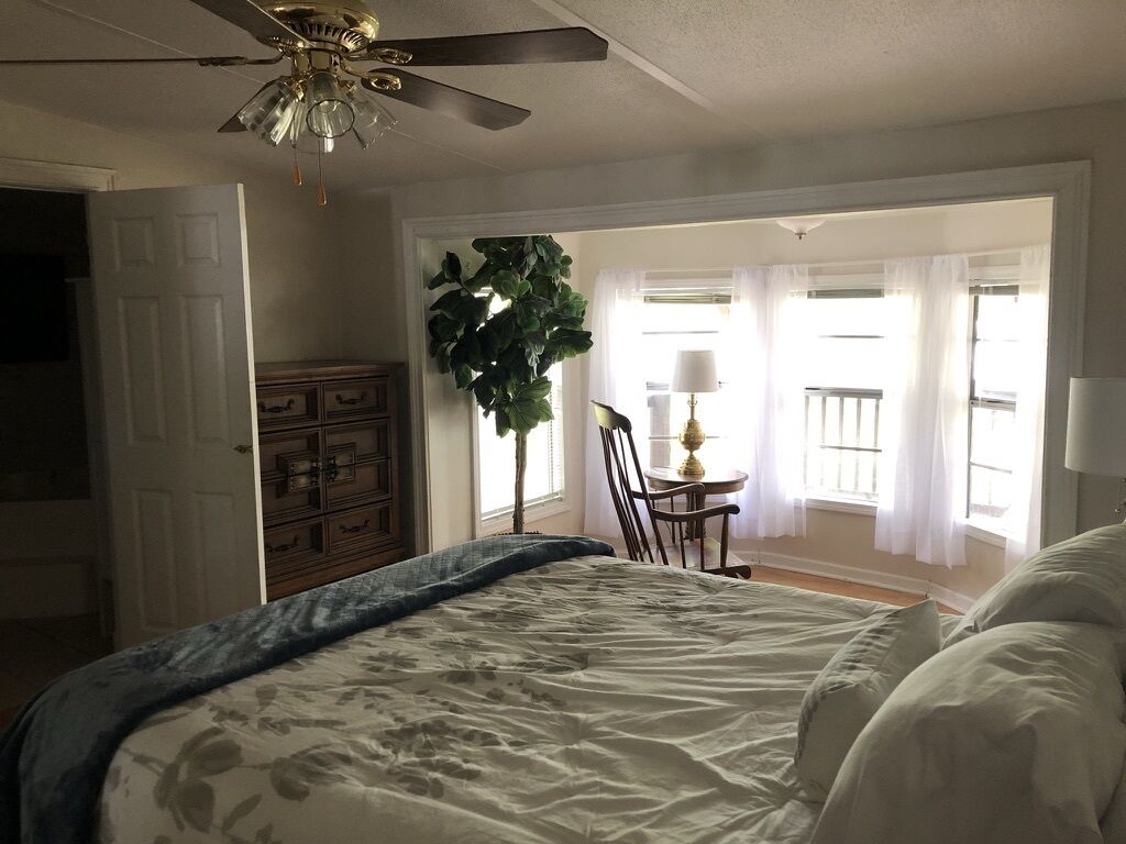 A bedroom with a bed, a ceiling fan, a dresser, a chair, a table, and a window with curtains.