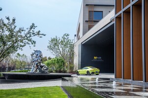 Un edificio moderno con fachada de vidrio, una escultura en primer plano y un coche deportivo amarillo estacionado al frente.