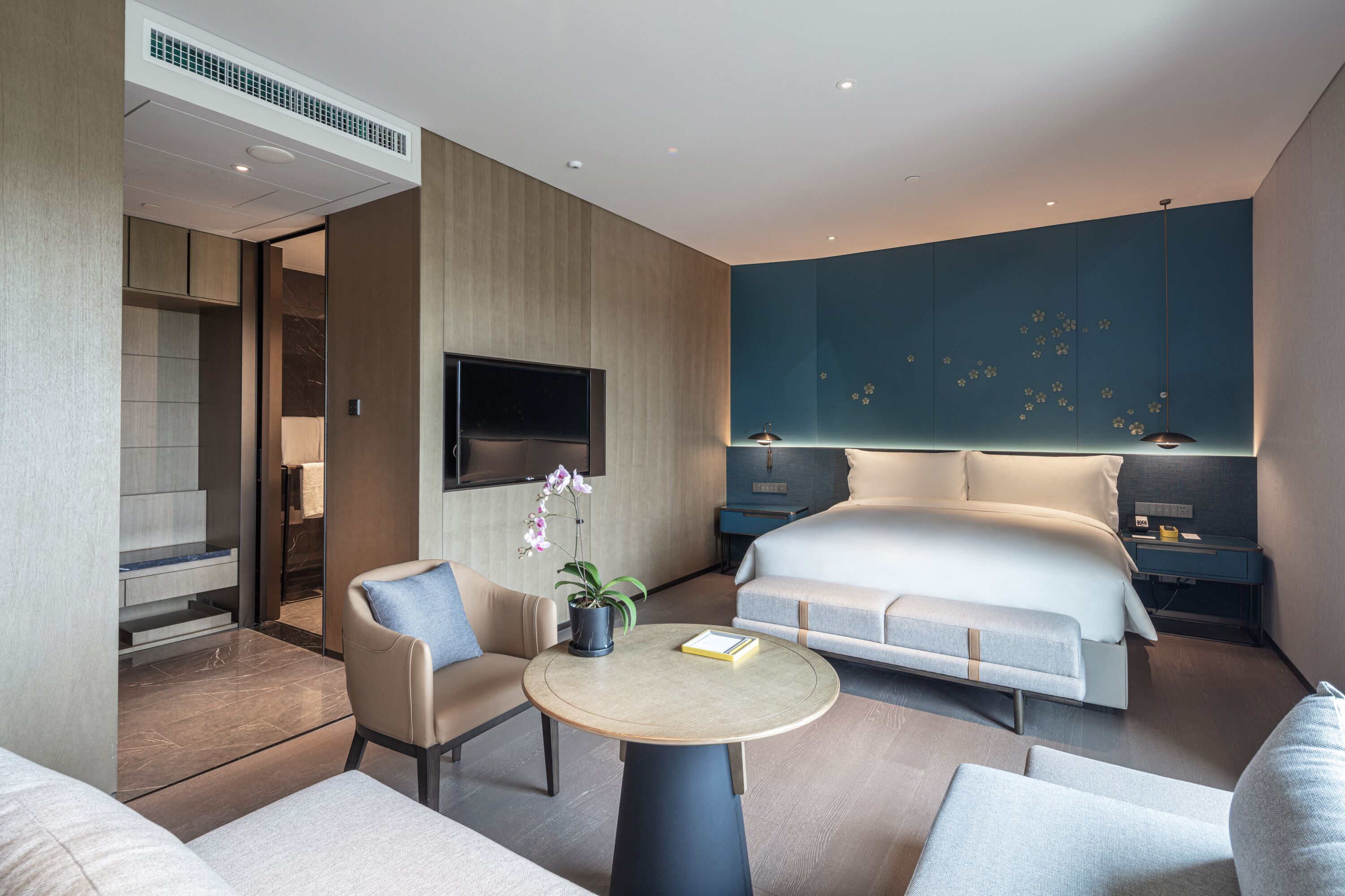 A modern hotel room with a large bed, a seating area with a round table, a TV mounted on the wall, and a small potted plant.