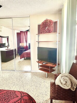Une chambre d’hôtel avec un lit, un téléviseur posé sur un meuble, un miroir et une fenêtre avec des rideaux.