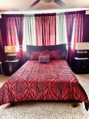 Un dormitorio con una cama grande, cortinas rojas y blancas, dos mesitas de noche con lámparas y un ventilador de techo.