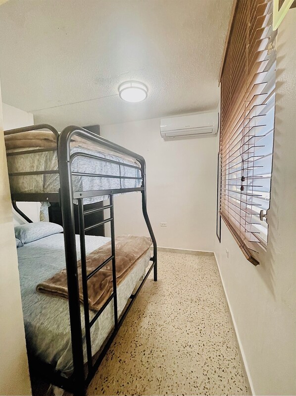 Une petite chambre avec un seul lit, un lit superposé, une fenêtre avec des stores, un appareil de climatisation et un sol à motifs.