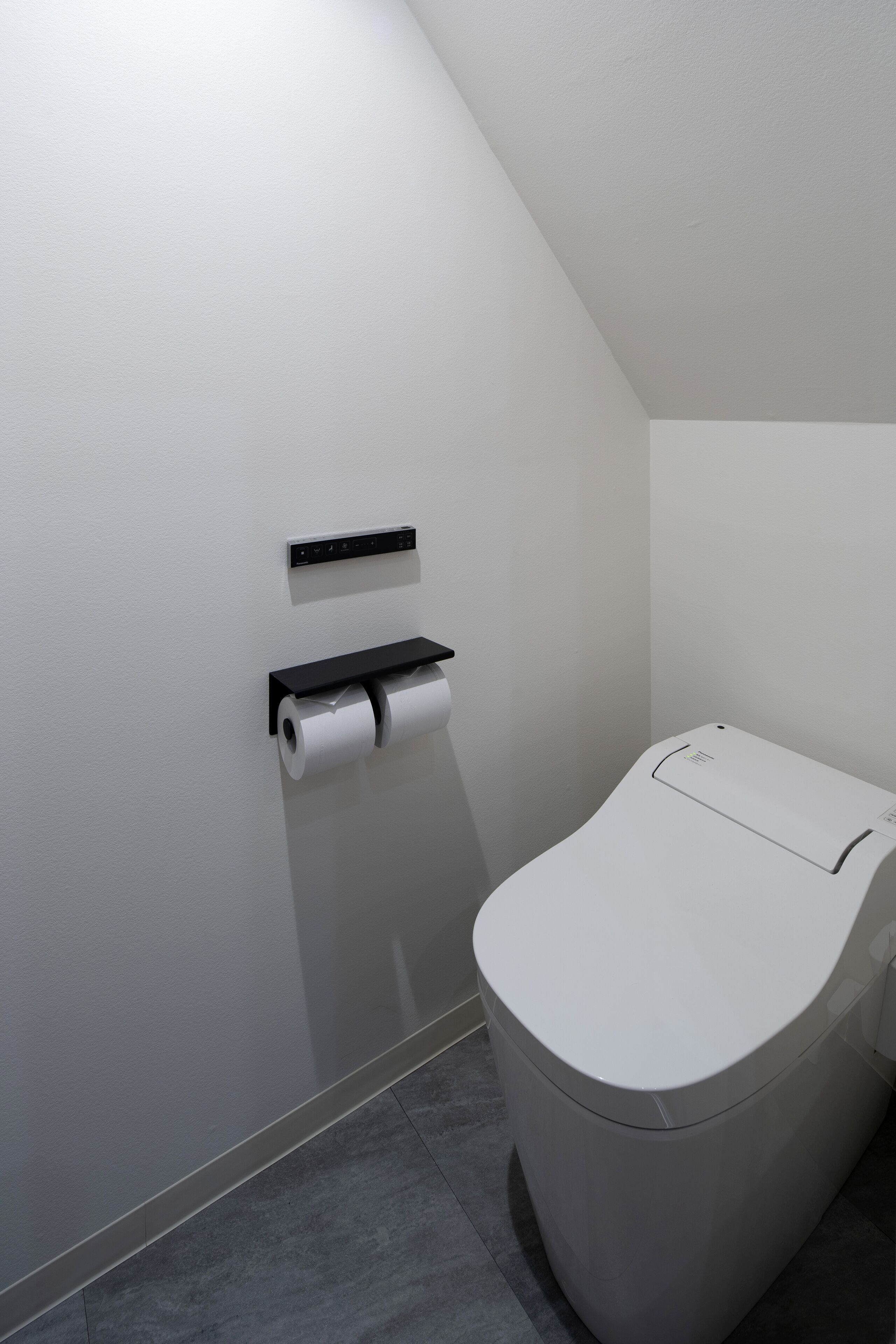 A modern bathroom with a toilet, a toilet paper holder, and a wall-mounted flush control panel.