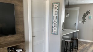 A modern living room with a wooden floor, a TV mounted on a wooden panel, a white door, a wall-mounted sign reading "BEACH", and a small bar area with stools.
