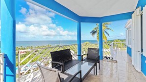 A balcony with a glass table and chairs, overlooking a coastal view with palm trees and a clear sky. - Ocean View & walk 5 min to beach, Pool Table, Ping Pong, Arcade Games 5Bd-4 Bth (Vieques)