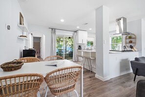 Une cuisine moderne avec des meubles blancs, un coin repas avec des chaises en osier et une vue sur la verdure à l’extérieur.