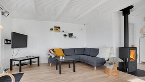 Una sala moderna con un sofá seccional gris, un televisor negro montado en la pared, una mesita, una silla de mimbre y un estufa de leña.