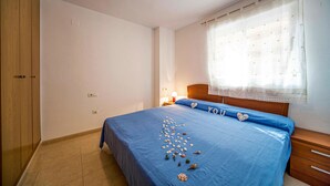 A bedroom with a blue bedspread, a wooden wardrobe, a nightstand with a lamp, and a window with sheer curtains.