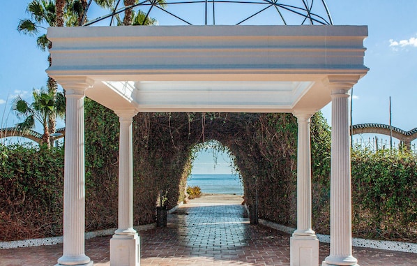 En vit pergola med pelare som leder fram till en plats med utsikt över stranden.