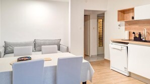 Uma sala de estar moderna e minimalista com uma mesa de jantar e cadeiras, uma cozinha com armários brancos e bancada de madeira, e um corredor que dá para outro quarto.