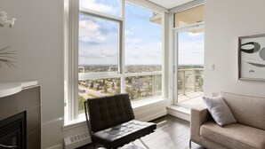 A modern living room with a large window offering a city view, a black leather chair, a beige sofa, and a framed abstract artwork on the wall. - Newly Furnished 2BD, 2BA Condo with Pool and Stunning View! (Calgary)