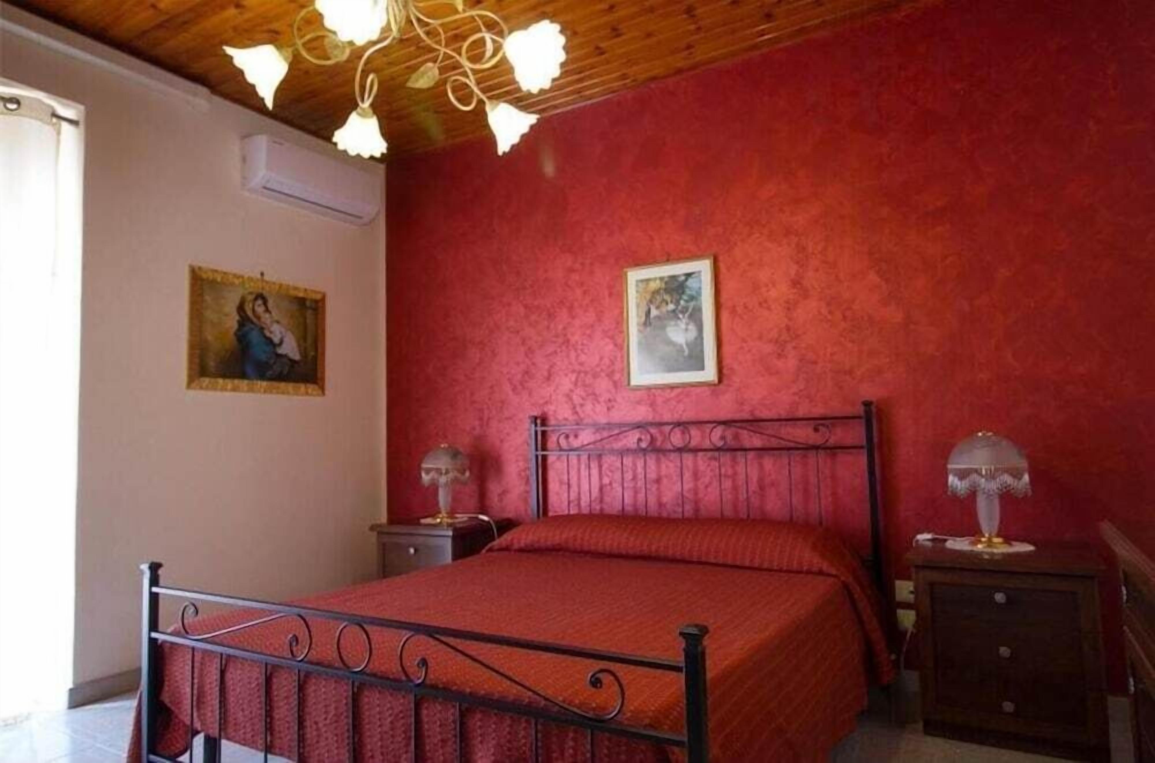 Un dormitorio con una pared roja, estructura de cama metálica, dos mesitas de noche con lámparas y un aplique de techo.