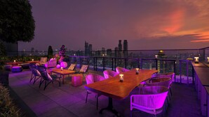 An outdoor terrace with a city skyline view, featuring a wooden table and chairs, and ambient lighting.
