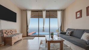 A living room with a sofa, a wooden bench, a coffee table, and a view of the sea through large glass doors. - Holiday Home ‘Casa Santa Úrsula’ with Sea View, Pool, Wi-Fi & Garden (Santa ursula)