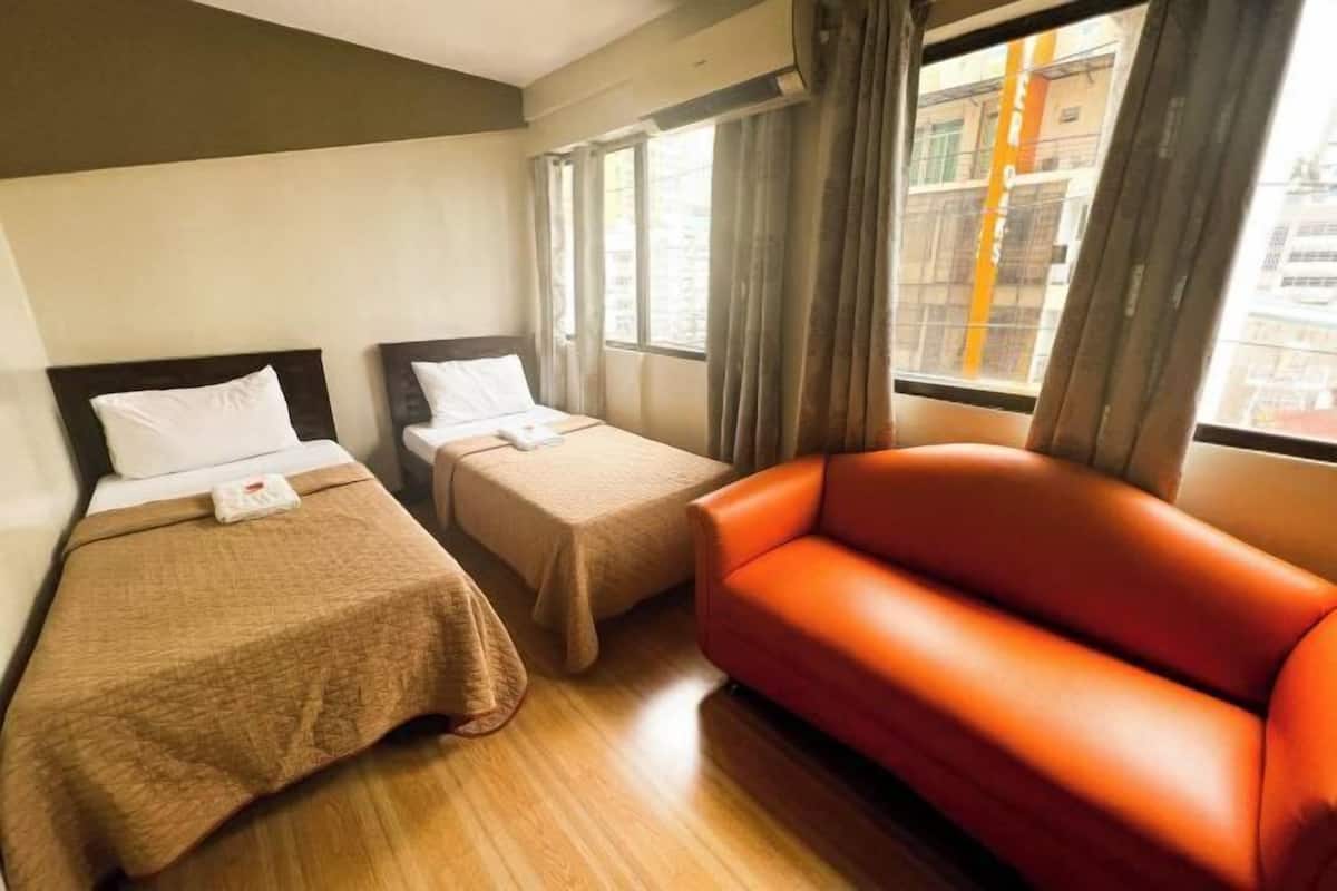A hotel room with two single beds, a red sofa, and a window with curtains.