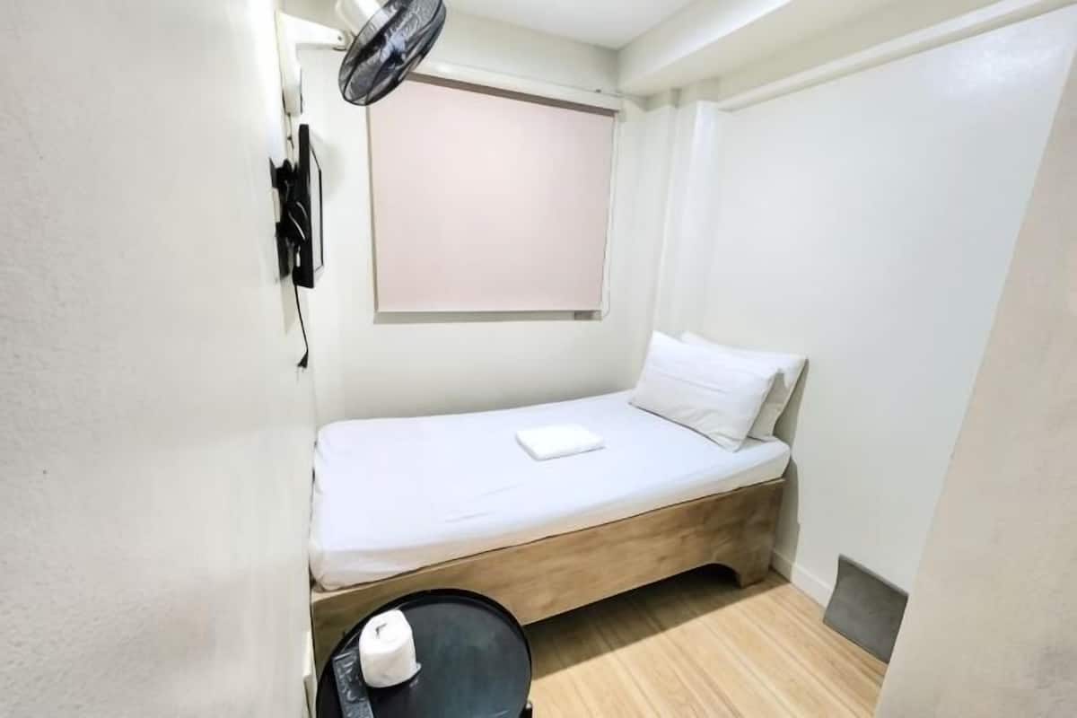 A small, single-bed room with a wooden floor, a window with a blind, a wall-mounted fan, and a small table with a white object on it.