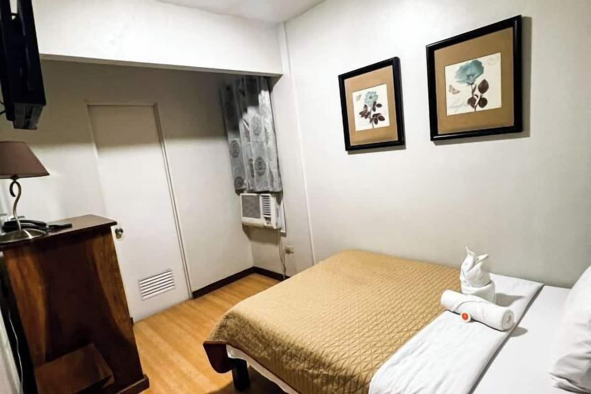A hotel room with a bed, a desk, a chair, a window with curtains, and two framed pictures on the wall.