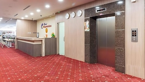 A hotel reception area with a reception desk, a clock, an elevator, and a red carpeted floor.