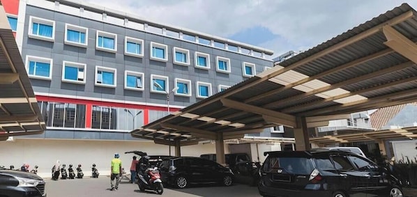 A covered parking area with multiple cars and motorcycles, a building with a modern facade, and a clear sky. - Grand Impression Hotel Medan (Medan)