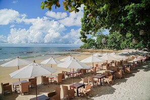 Un restaurante frente a la playa con terraza, sombrillas blancas y mesas.