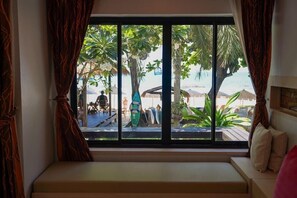 Una habitación con un ventanal grande que da a una playa, una banca y vistas a palmeras y sombrillas de playa.