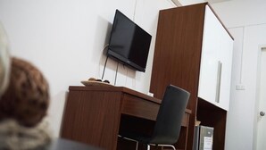 A room with a wooden desk, a black chair, a television mounted on the wall, and a small refrigerator.