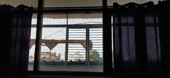 A window with a metal grid, dark curtains, and a view of a building with a dome and trees. - Joey's Hostel (Agra)