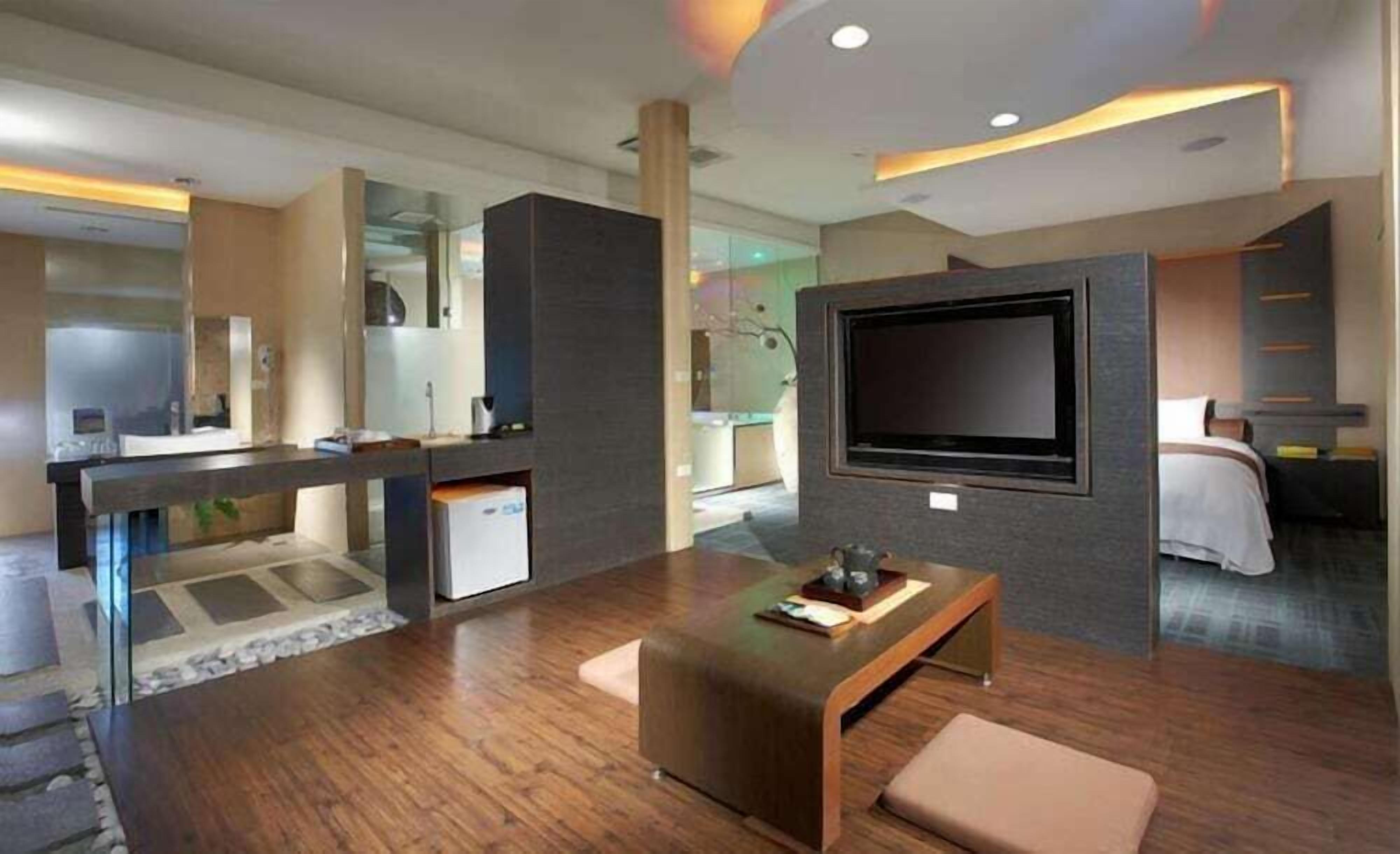 Ein modernes Hotelzimmer mit großem Flachbildfernseher an der Wand, Holzboden, einer Sitzecke mit einer Bank und einem großen Spiegel.