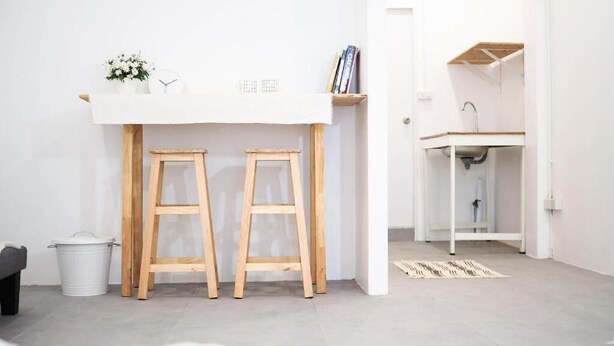 A minimalist room with a white desk, two wooden stools, a small white bucket, and a white cabinet with a sink. - Unyak Room Ayutthaya (Ayutthaya)