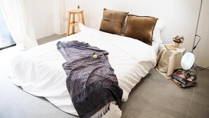 A neatly made bed with a white and brown blanket, a small wooden nightstand with a lamp, and a stack of books. - Unyak Room Ayutthaya (Ayutthaya)