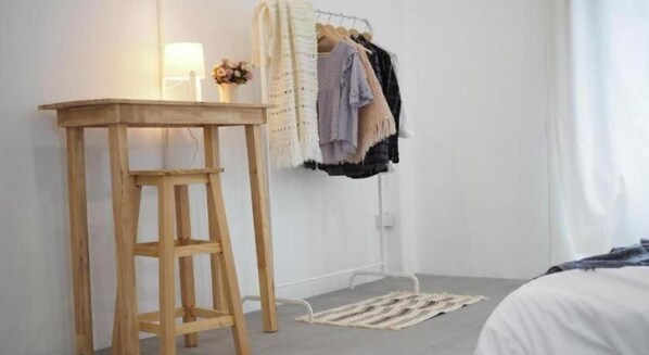 A room with a wooden stool, a clothing rack with various garments, a lamp, and a bed with a patterned rug.