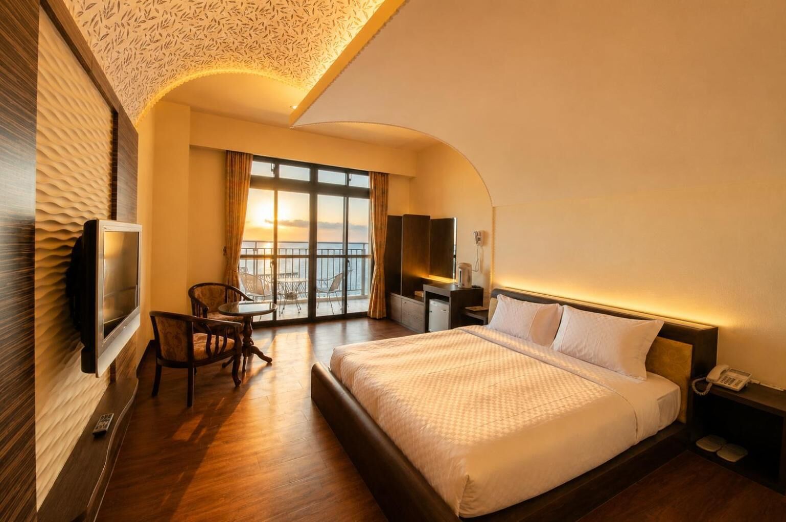 A hotel room with a large bed, a desk with a chair, a television, and a balcony with a view.