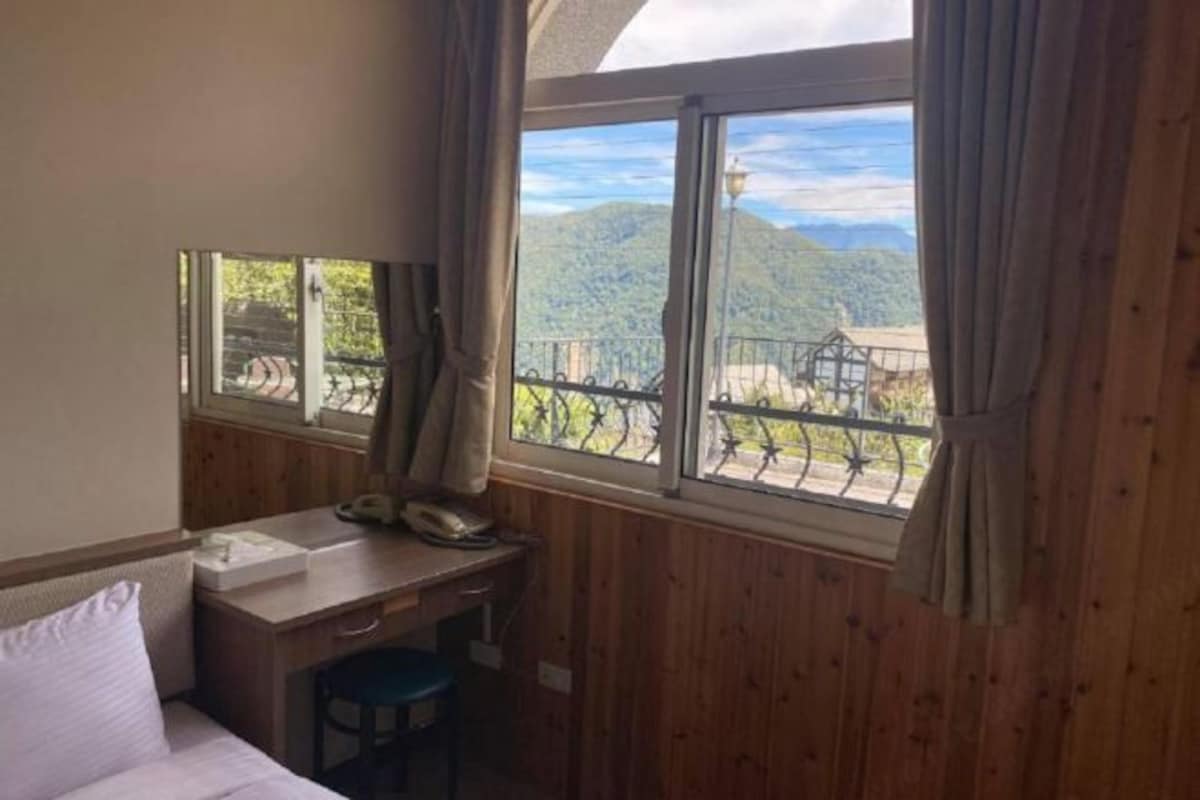 A room with a wooden desk, a bed, and a window with a view of mountains and a fence.