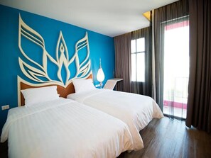 A hotel room with two beds, a blue accent wall, a large window with curtains, and a nightstand with a lamp. - Vassana Design Hotel (Ayutthaya)