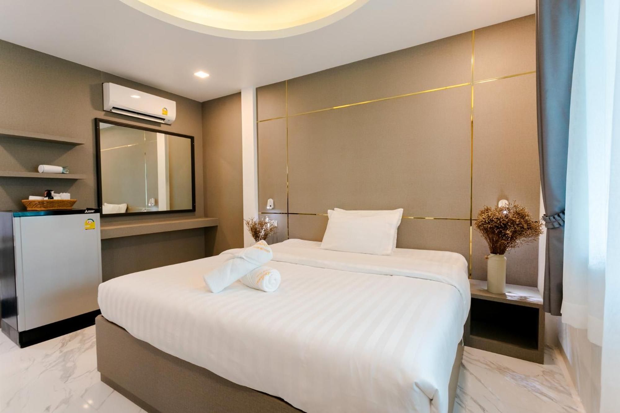 A hotel room with a bed, a mirror, an air conditioning unit, a small refrigerator, and a vase with dried flowers.