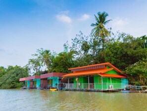 A floating house with a green and orange roof, surrounded by water and palm trees. - The Neenlawat Riverside (Phunphin)
