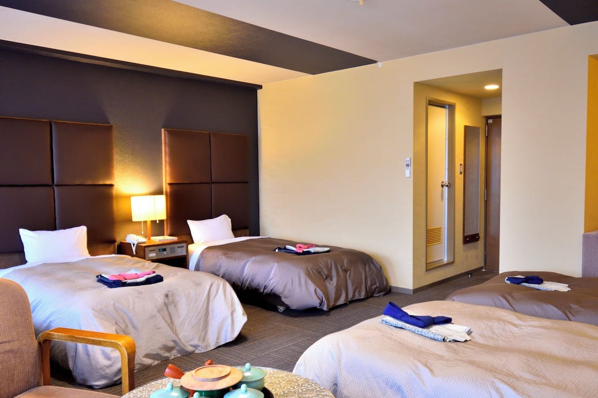 Ein Hotelzimmer mit drei Betten, einem Sessel, einem Nachttisch mit Lampe und einem Spiegel.